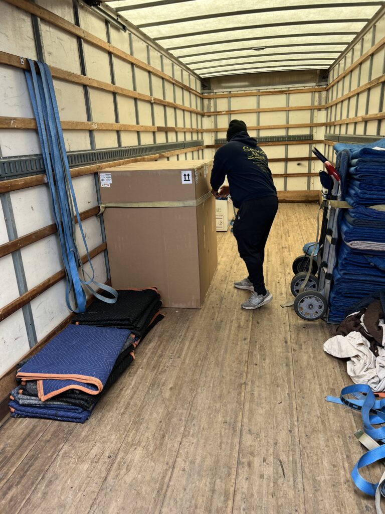 A person arranges a large box inside a moving truck, with moving blankets and equipment neatly stacked nearby, preparing for transport.