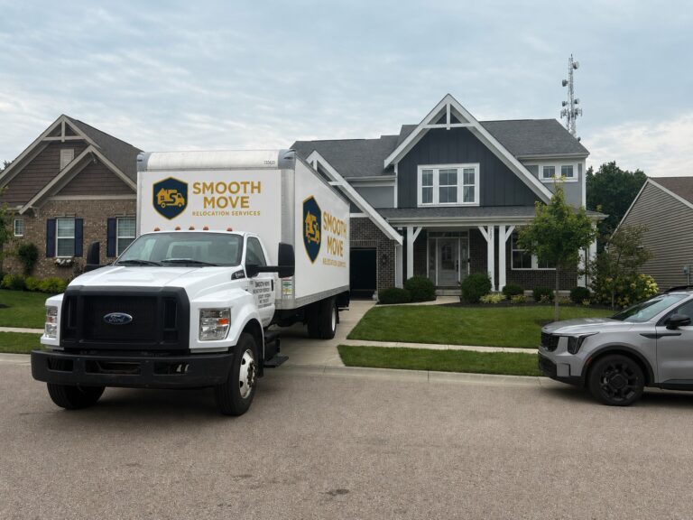 White moving truck with Smooth Move Relocation Services parked in a suburban street in front of a gray-blue house with a manicured lawn.